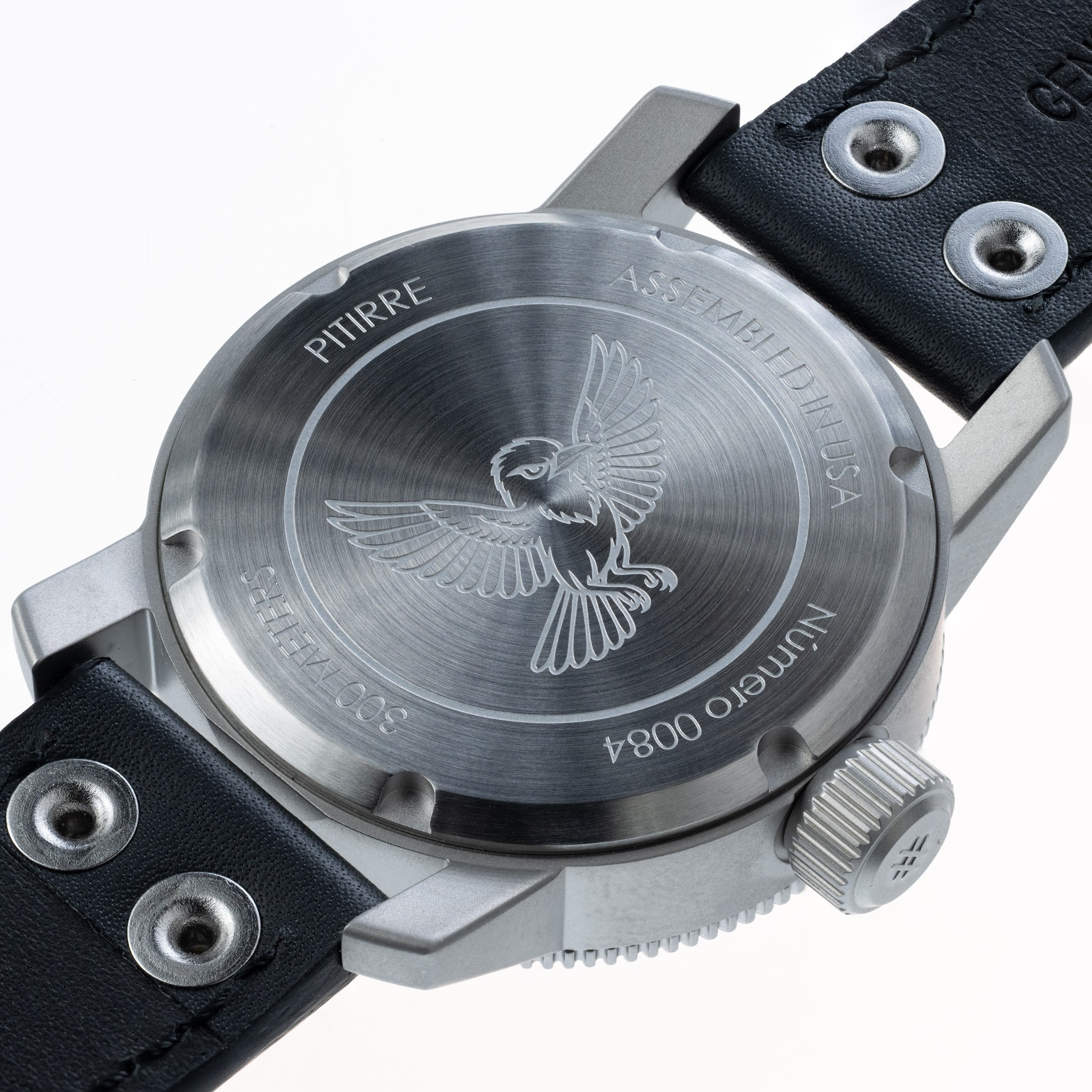 Close-up of the back of a wristwatch with a brushed silver metal case and a black leather strap. The case is engraved with a bird motif and text including 'PITIRRE' and 'ASSEMBLY USA'.