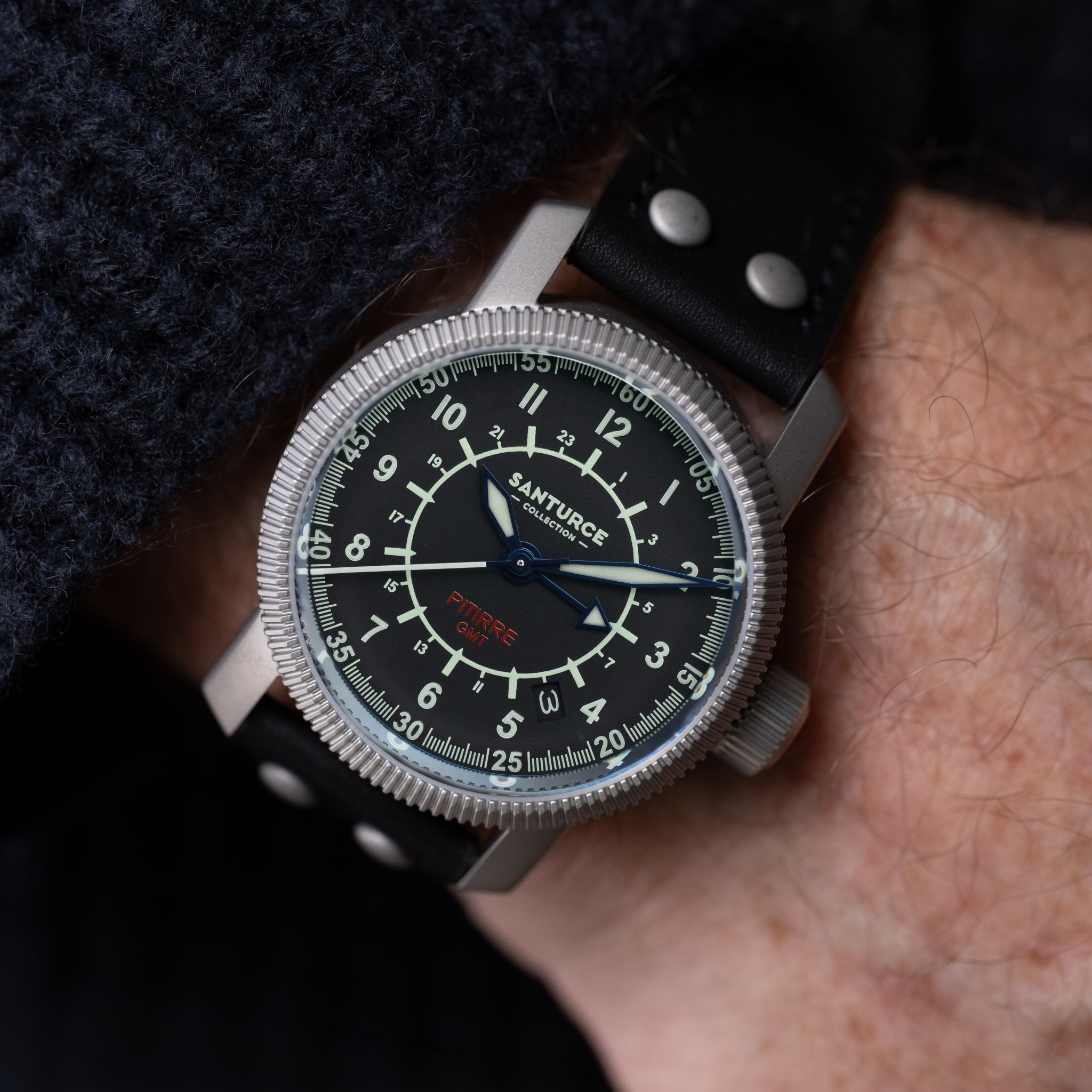 A close-up of a wrist wearing a Santurce Pitirre GMT watch with a black dial, silver-tone case, and black leather strap. The watch brand 'SANTURCE' is visible on the dial.