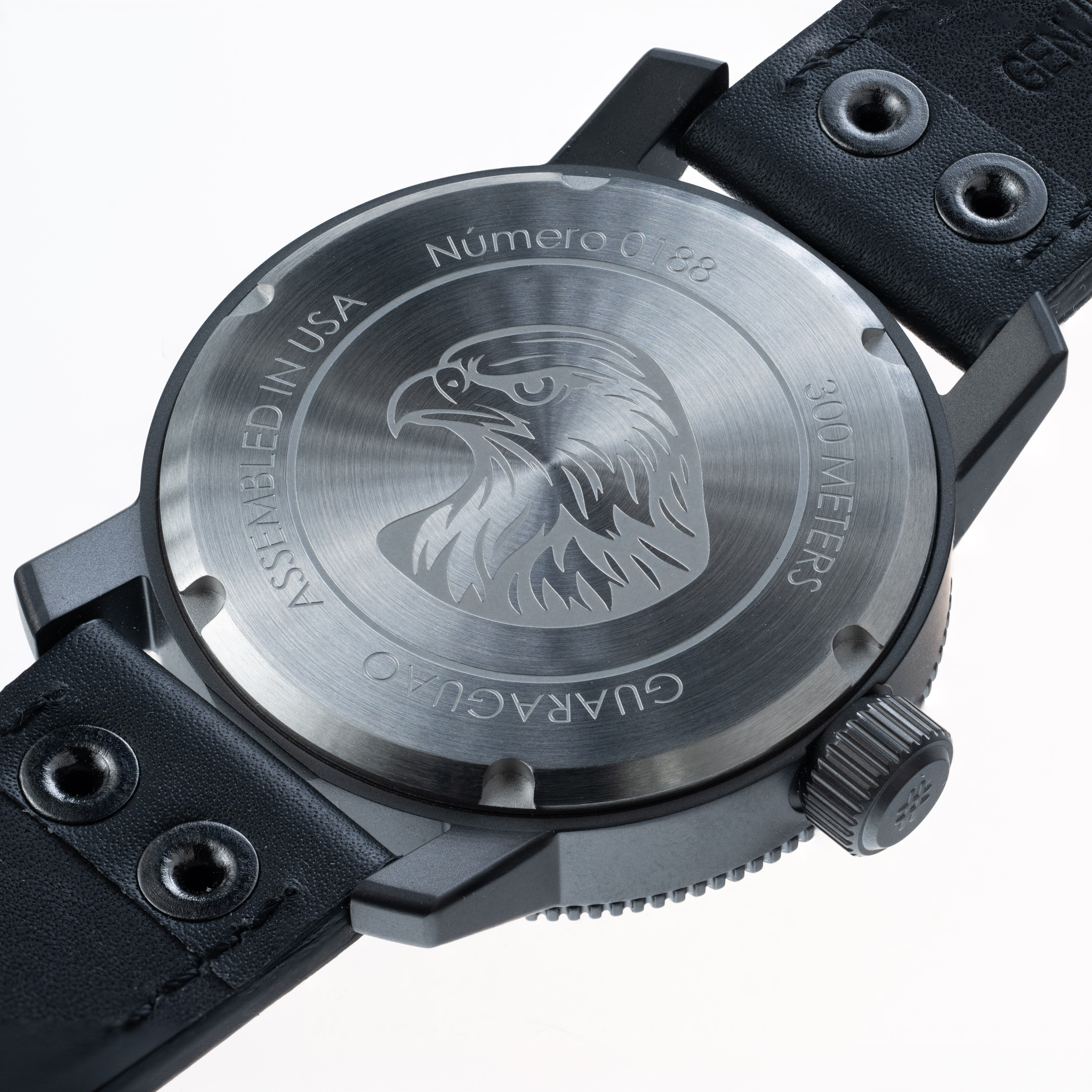 Close-up of the back case Guaraguao wristwatch with a brushed silver metal case back engraved with an eagle head and text, attached to a black leather strap on a white background.